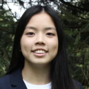 Jasmine Gong smiling confidently in a blazer, standing outdoors among green pine trees, reflecting her leadership and martial arts mastery.
