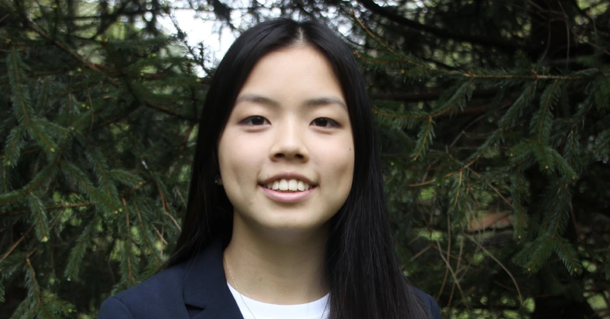 Jasmine Gong smiling confidently in a blazer, standing outdoors among green pine trees, reflecting her leadership and martial arts mastery.