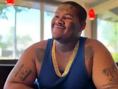 Crip Mac smiling in a restaurant, showcasing his tattoos and gold chain, representing his success and lifestyle for Crip Mac Net Worth.