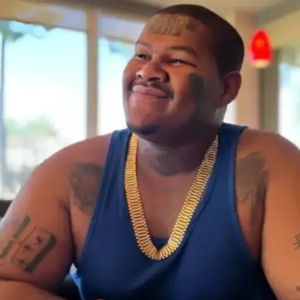 Crip Mac smiling in a restaurant, showcasing his tattoos and gold chain, representing his success and lifestyle for Crip Mac Net Worth.
