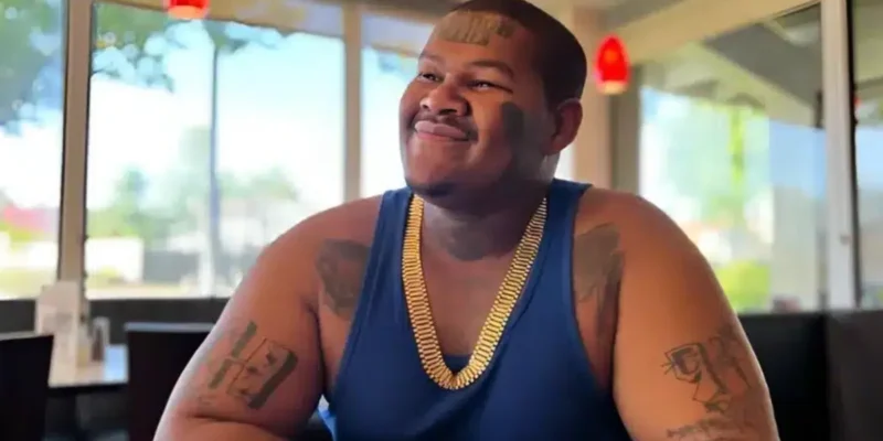 Crip Mac smiling in a restaurant, showcasing his tattoos and gold chain, representing his success and lifestyle for Crip Mac Net Worth.