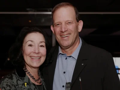 Gal Tirosh smiling with a woman at a formal event, both dressed elegantly and posing together in a warmly lit indoor setting.