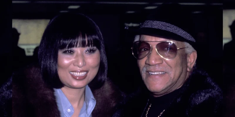 Kaho Cho smiling alongside comedy legend Redd Foxx at their wedding celebration in formal attire.