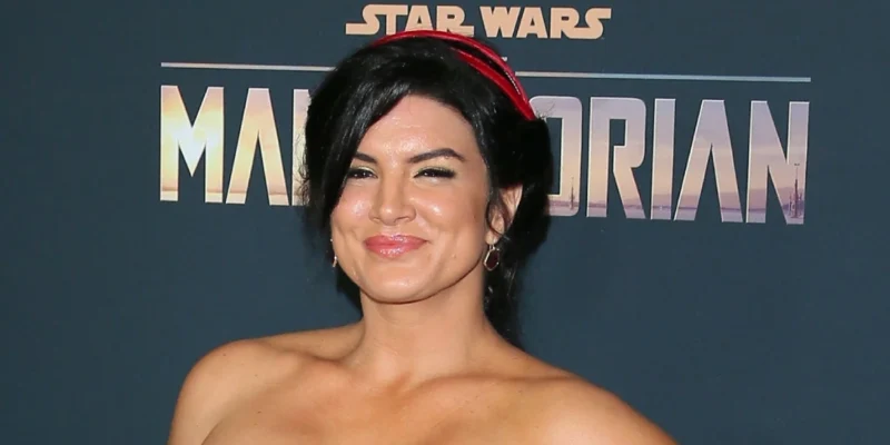 Gina Carano at The Mandalorian premiere, wearing a strapless dress and red headband, standing in front of the Star Wars backdrop.